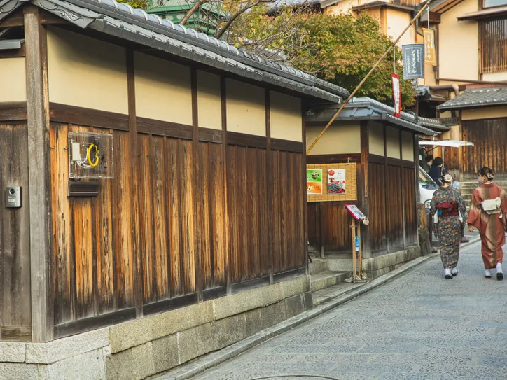 Back view of unrecognizable local Japanese ladies in traditional kimonos walking on paved street near typical aged wooden houses in Kyoto
