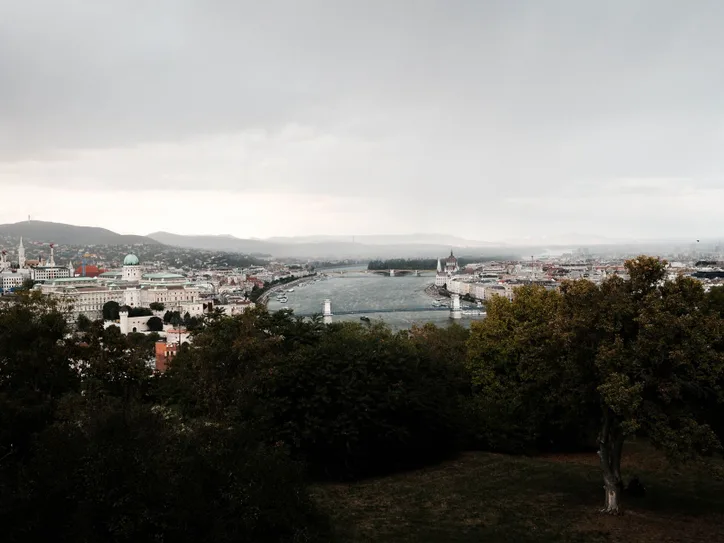 Scenic view of Budapest's cityscape with the Danube River and iconic landmarks under a cloudy sky.