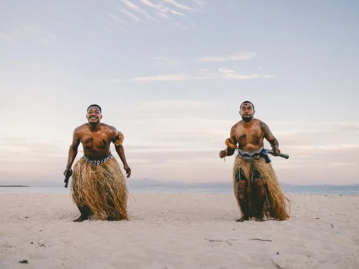Two traditional Fijian warriors perform on a sandy beach in Nadi, Fiji, at sunset.