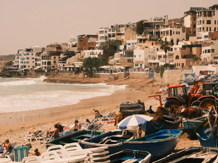 Scenic view of Taghazout beach with colorful boats, sunbathers, and adjacent hillside buildings.