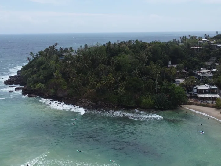 Drone shot capturing the beautiful coastline of Hiriketiya Beach, Sri Lanka, with lush greenery and clear blue waters.